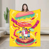 A yellow throw blanket featuring a playful cartoon illustration of a hamburger and a family standing on the bun.