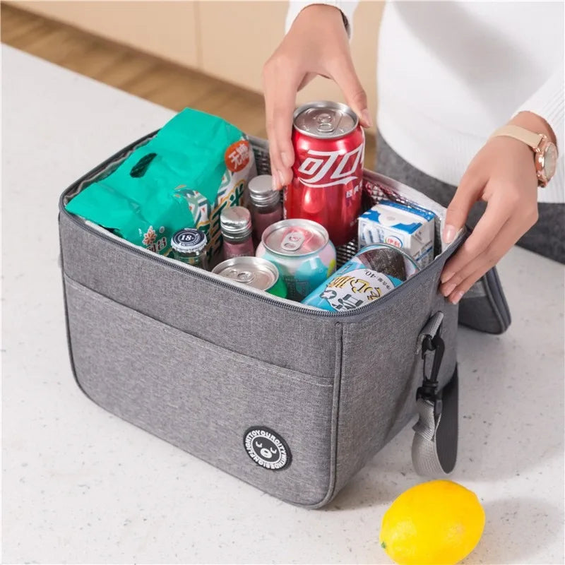 A hand is placing a can of soda into a gray thermal insulated lunch bag on a white surface.
