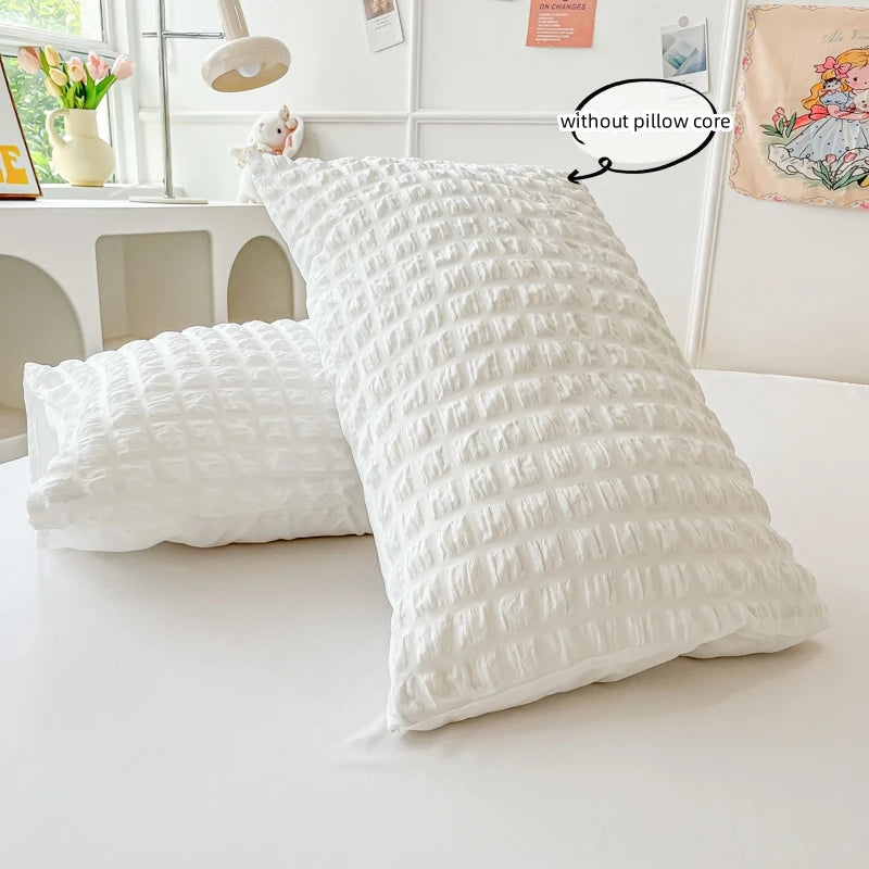 A white, textured pillow is shown next to a fluffy white pillow on a white surface.