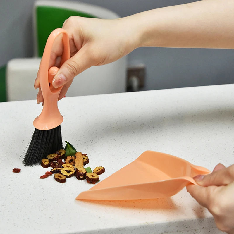 A hand is holding an orange compact mini broom and dustpan set on a white desk surface.