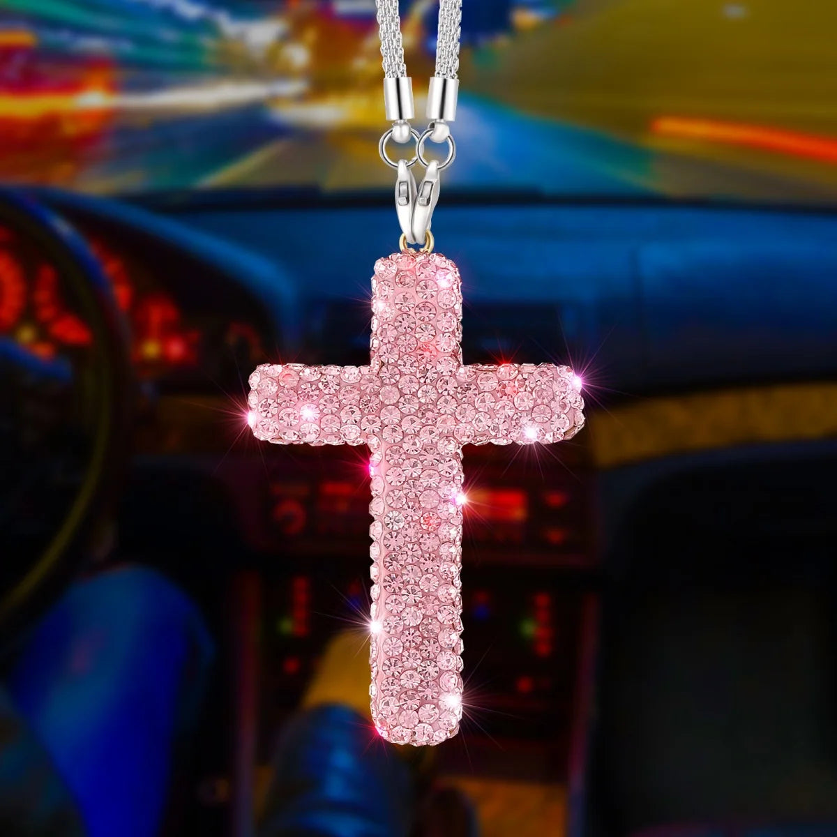 A pink rhinestone cross pendant suspended by a silver chain, displayed against a blurred car interior background.