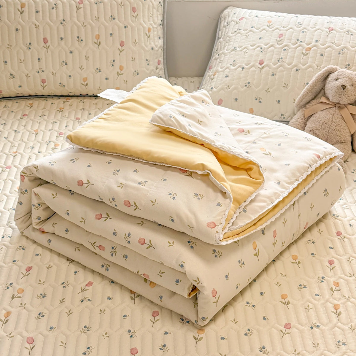 A stack of cream and yellow lace-trimmed breathable summer duvets resting on a quilted bedspread.