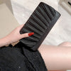 A hand holds a black pleated clutch bag with a chain strap against a light background.