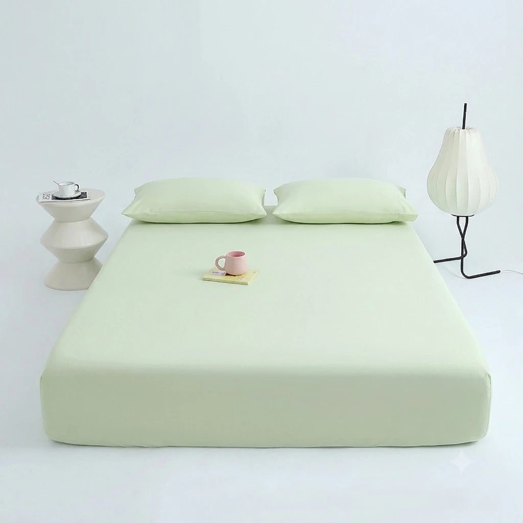 A light green cooling ice-silk fitted sheet is displayed on a white surface with a small pink cup and a glass lamp in the bac