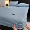 A light blue plush mattress cover with an elastic edge is shown on a bed, being held by a hand.