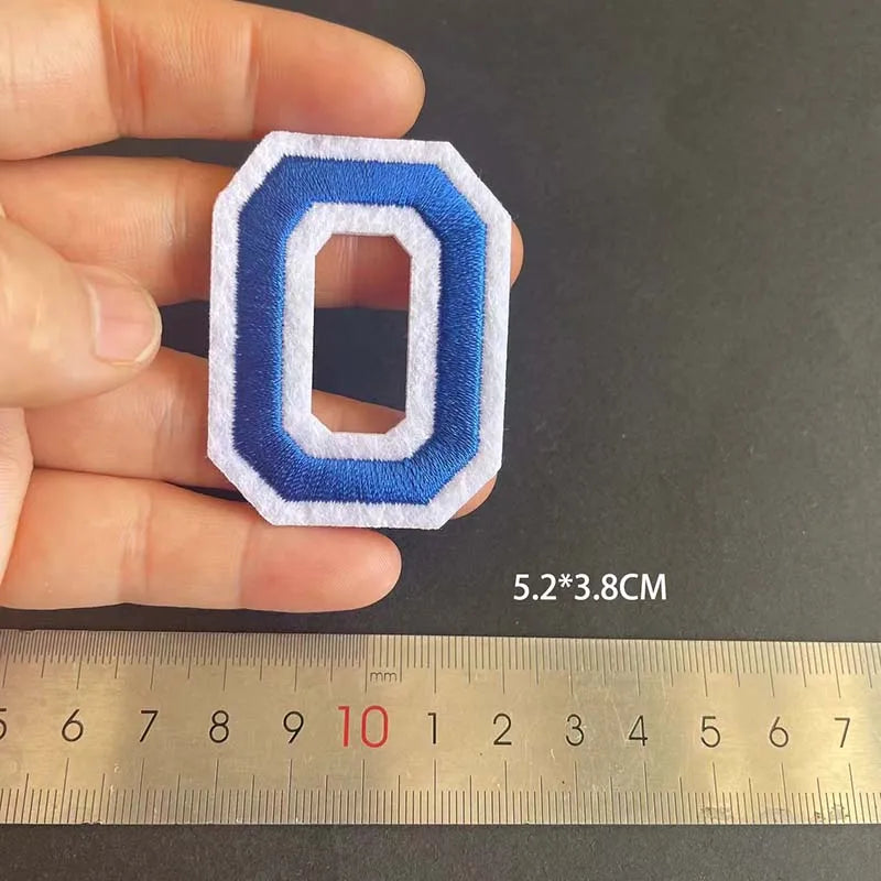 A hand holds a blue and white embroidered letter O patch, measuring 5.2 by 3.8 cm against a ruler.