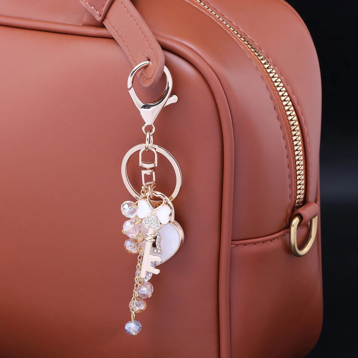 A gold-toned keychain charm featuring a rhinestone bow and heart attached to a leather handbag strap.