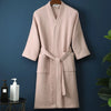 A light pink waffle knit robe hanging on a wooden hanger against a dark blue wall.