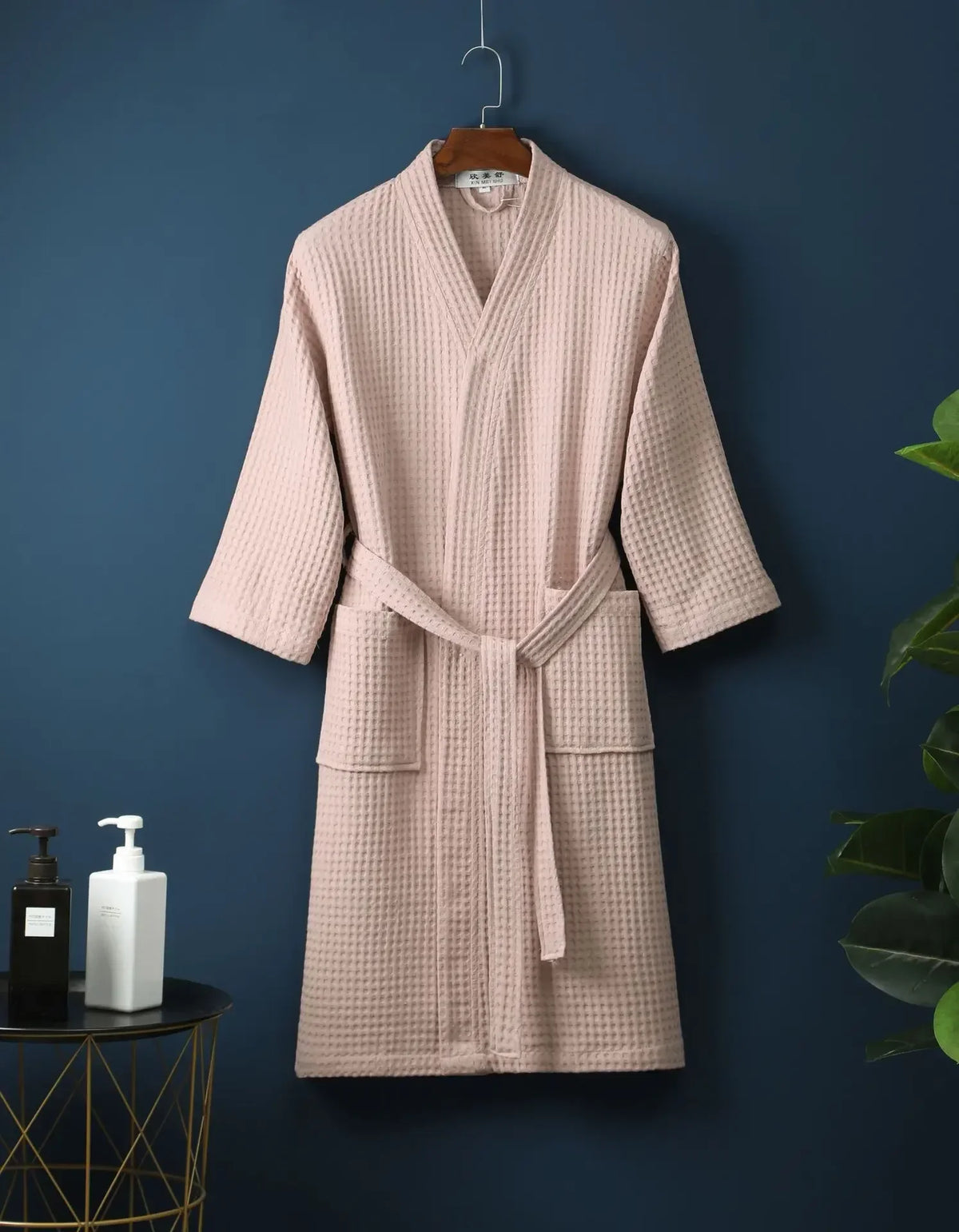 A light pink waffle knit robe hanging on a wooden hanger against a dark blue wall.