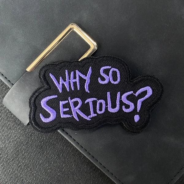 A purple embroidered patch with the text Why So Serious is attached to a black leather item.