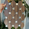 A hand holds a hexagonal mosaic tile panel made of dark brown wood and clear glass accents, displayed against green foliage.