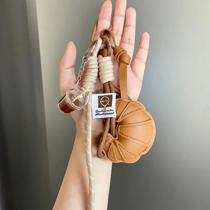 A hand holds a braided leather croissant keychain with a bag pendant and attached to a light-colored cord.