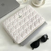 A quilted white laptop sleeve with a pink floral pattern is shown next to a silver laptop and a glass perfume bottle.