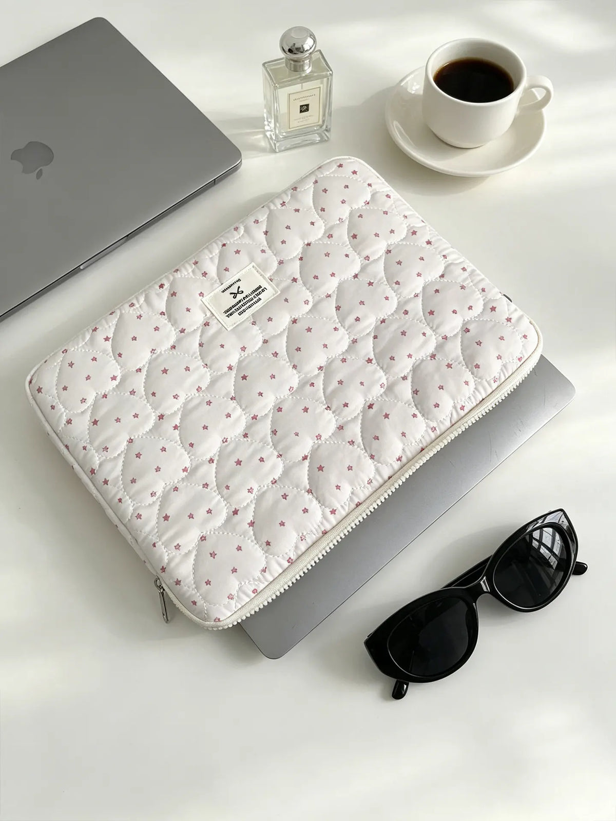A quilted white laptop sleeve with a pink floral pattern is shown next to a silver laptop and a glass perfume bottle.