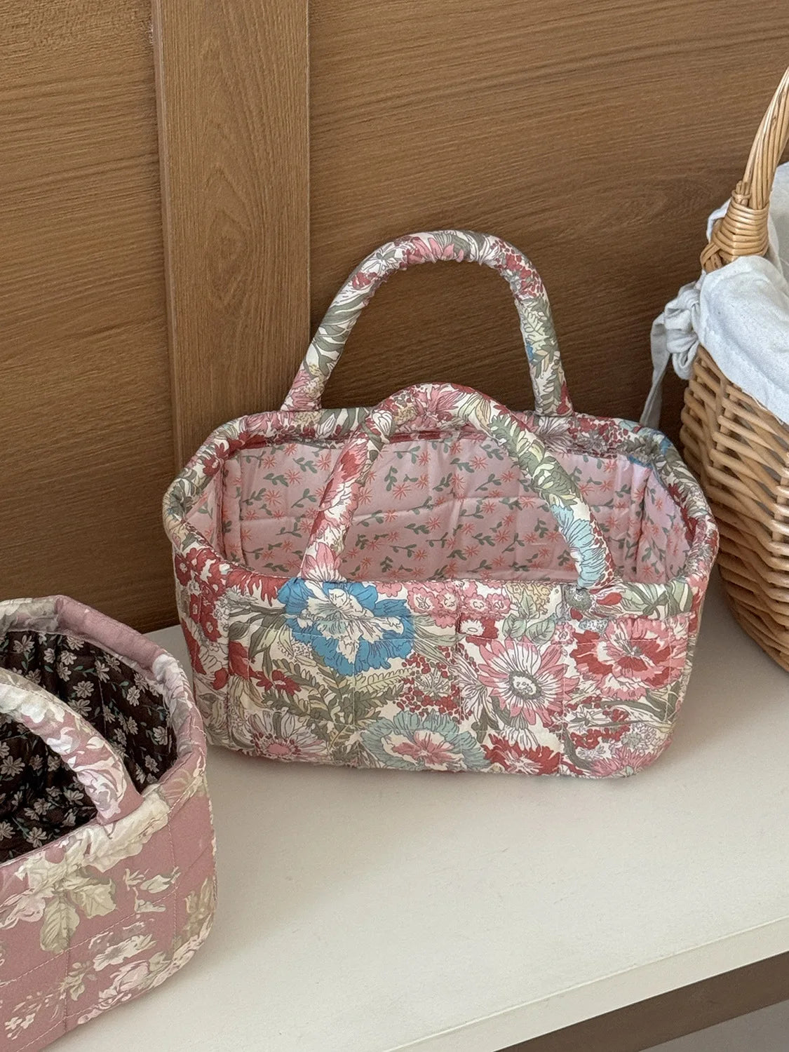 A pink quilted fabric storage basket with a handle and floral pattern sits next to another similar basket.