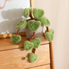 A small potted plant made of soft green faux leaves resembling a pothos, sitting on a wooden surface.