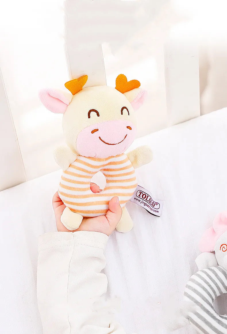 A hand holds a soft plush baby rattle toy shaped like an orange and pink cartoon animal.