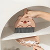 A hand is holding a brown cartoon bear broom and dustpan set against a white shelf.