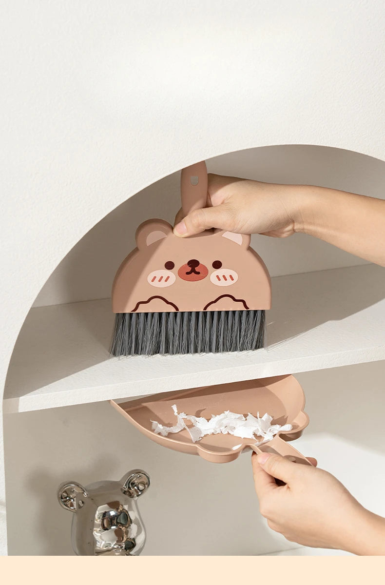 A hand is holding a brown cartoon bear broom and dustpan set against a white shelf.