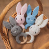A collection of crochet bunny rattles in pink, blue, and gray with natural wood teether rings resting in a woven basket.