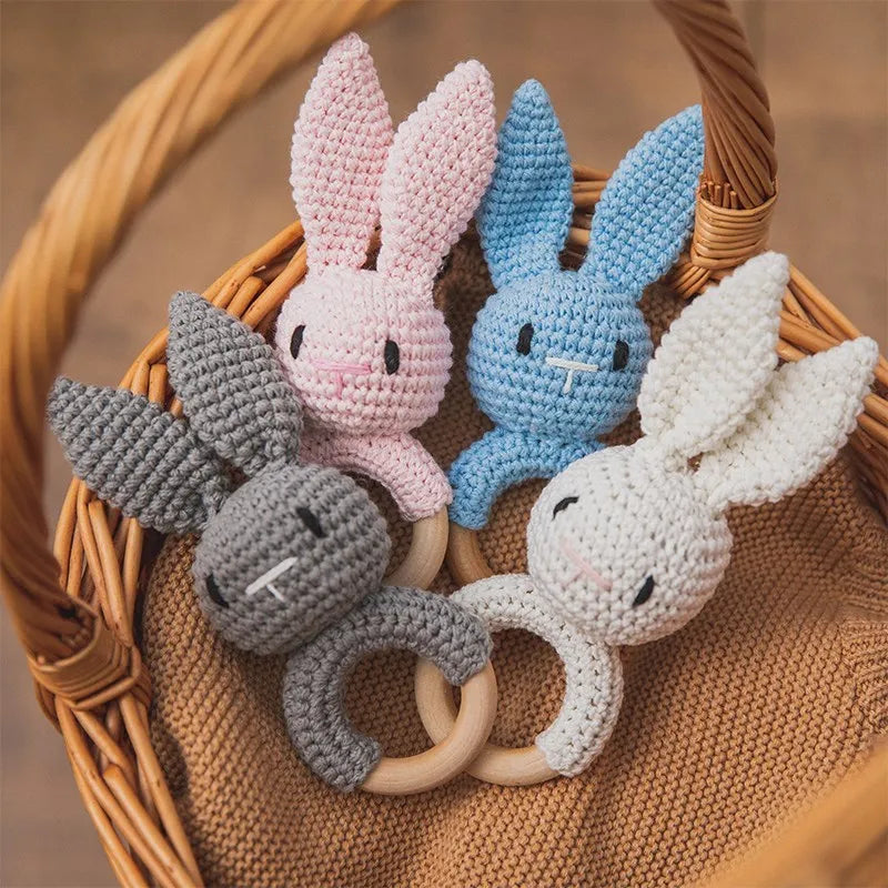 A collection of crochet bunny rattles in pink, blue, and gray with natural wood teether rings resting in a woven basket.