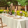 A long table set for a wedding or formal dinner with white satin tablecloths, candles, and floral arrangements.