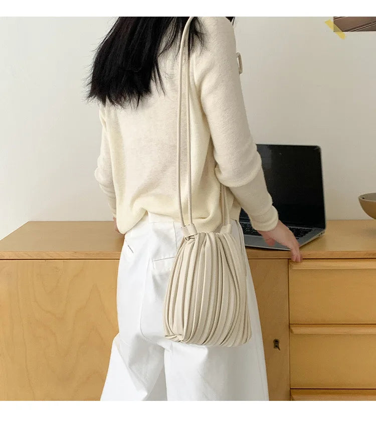 A woman wearing a cream sweater and white pants holds an elegant pleated faux leather crossbody bag.