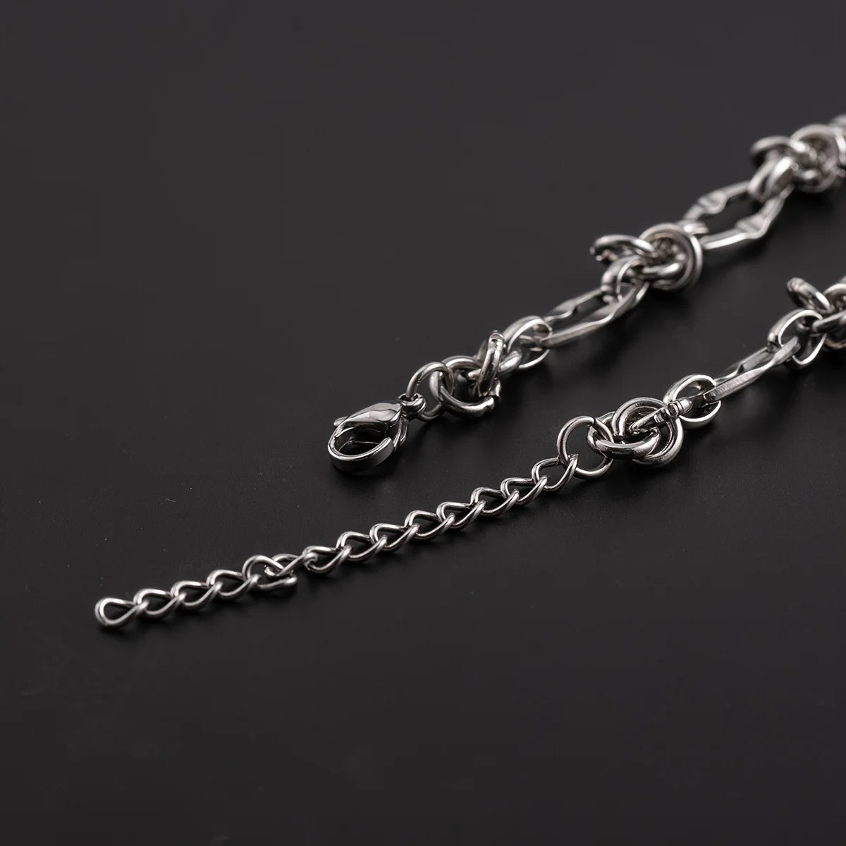 A silver titanium steel chain bracelet with knot details laid diagonally on a dark background.