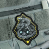 A black and yellow tactical snake morale patch with a hook-and-loop backing is shown on a gray fabric surface.