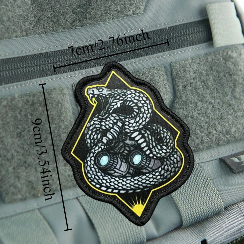 A black and yellow tactical snake morale patch with a hook-and-loop backing is shown on a gray fabric surface.