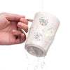A hand is pouring water into a ceramic mug decorated with an inspirational Bible verse sticker.