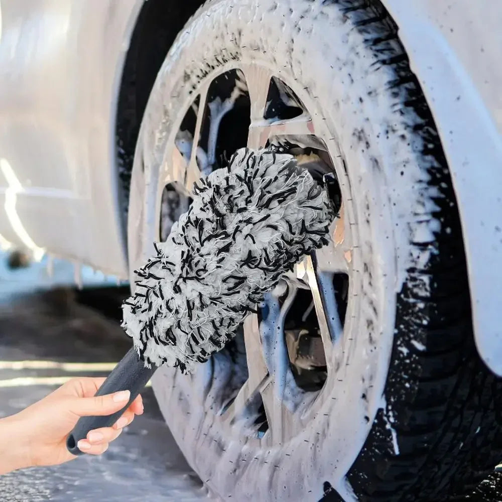 A hand is using a black brush to clean snow and dirt from a silver car wheel.