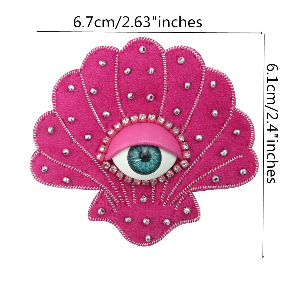 A pink fabric patch shaped like a flower with a central blue eye made of sparkling rhinestones.