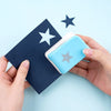 A hand holds a blue embossing punch next to a dark blue stamp on a light blue background.