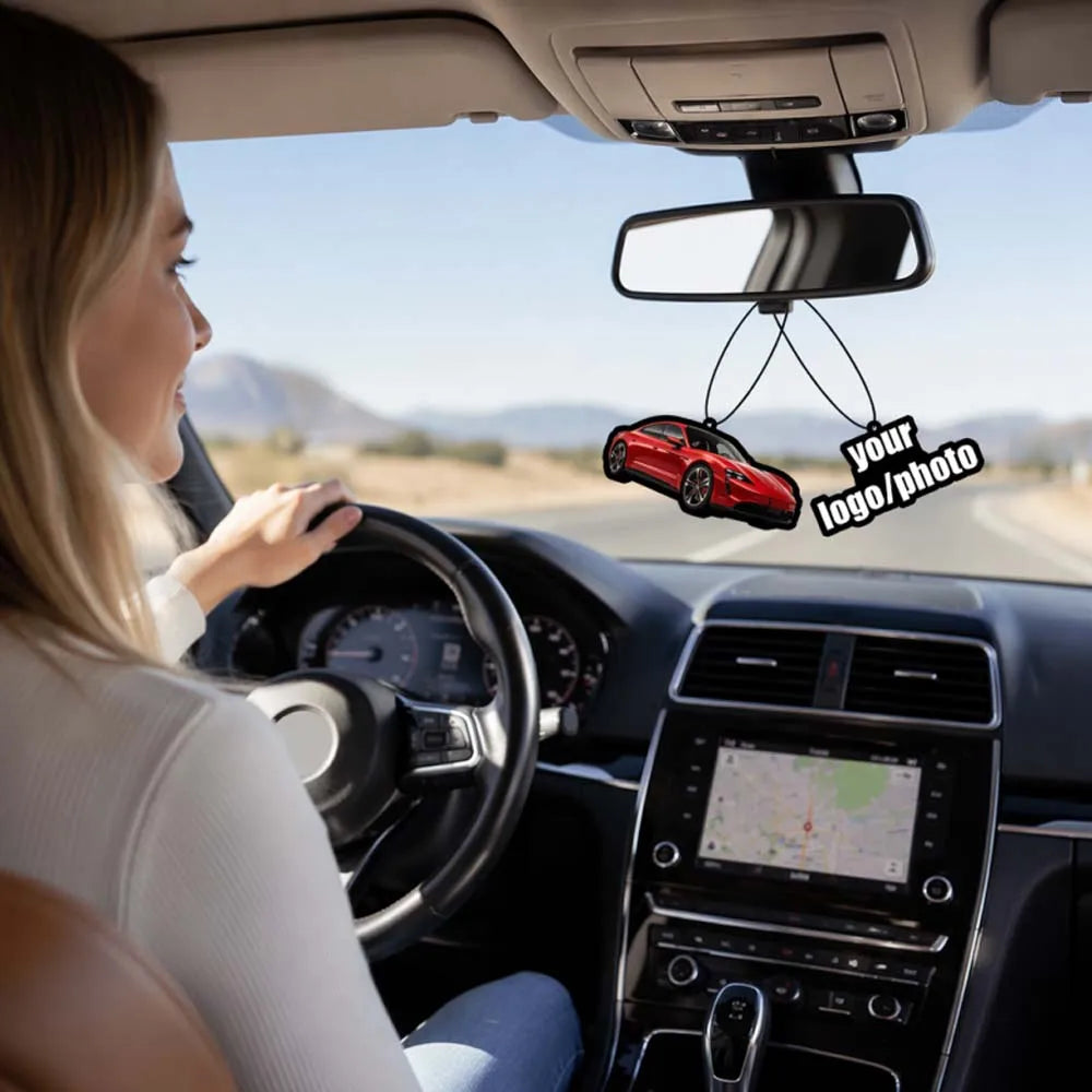 A woman drives a car with a custom photo air freshener hanging from the rearview mirror.