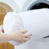 A hand holds a white mesh shoe washing bag with a zipper closure, positioned near a washing machine.