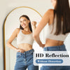 A woman is looking in a full-length mirror showing an HD reflection of herself wearing a white top and blue jeans.
