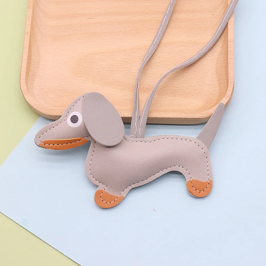 A tan dachshund-shaped PU leather keychain with an ear tag and leather collar rests on a light wooden tray.