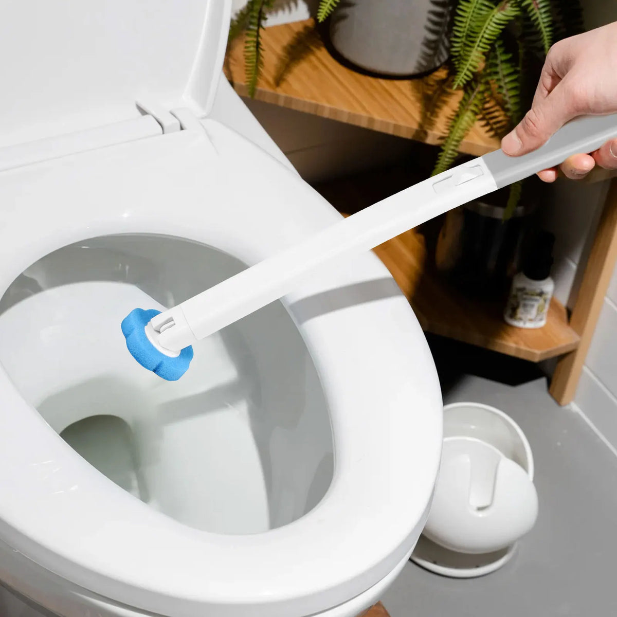 Une main tient une brosse de toilette blanche jetable avec une longue poignée au-dessus d'une cuvette de toilette blanche.