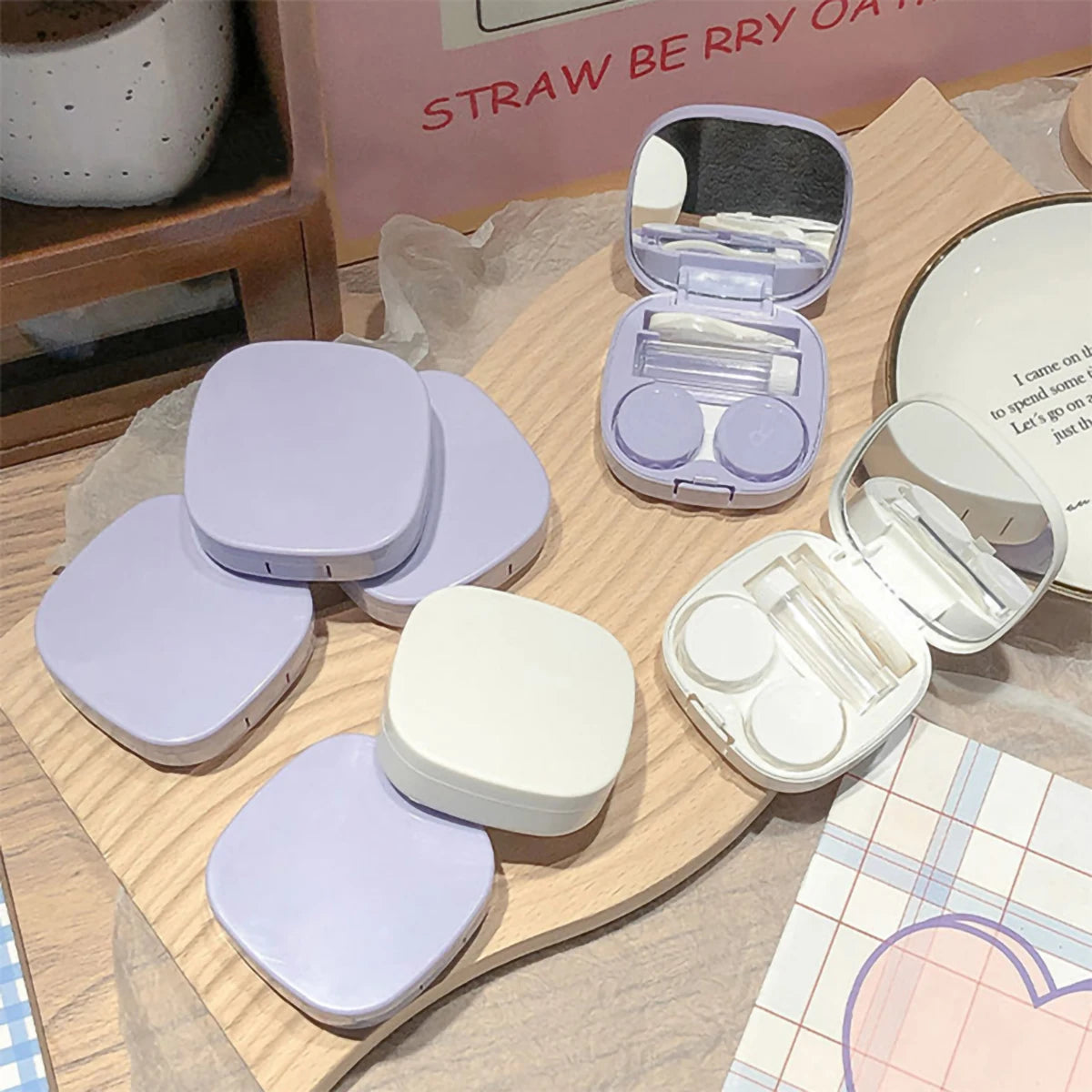A collection of purple and white compact contact lens travel cases and a mirror are displayed on a wooden surface.