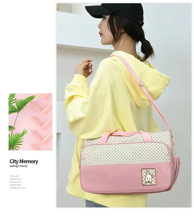 A woman wearing a yellow hoodie holds a pink and white patterned multifunctional crossbody diaper bag.