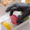 A hand in a black nitrile glove is holding and arranging colorful macarons inside a white box.