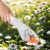 A hand is holding a white handheld insect catcher clamp tool in a sunny garden filled with white and yellow flowers.