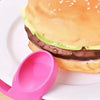 A sandwich made with a golden bun, dark spread, and green filling, served on a white plate next to a pink silicone tool.