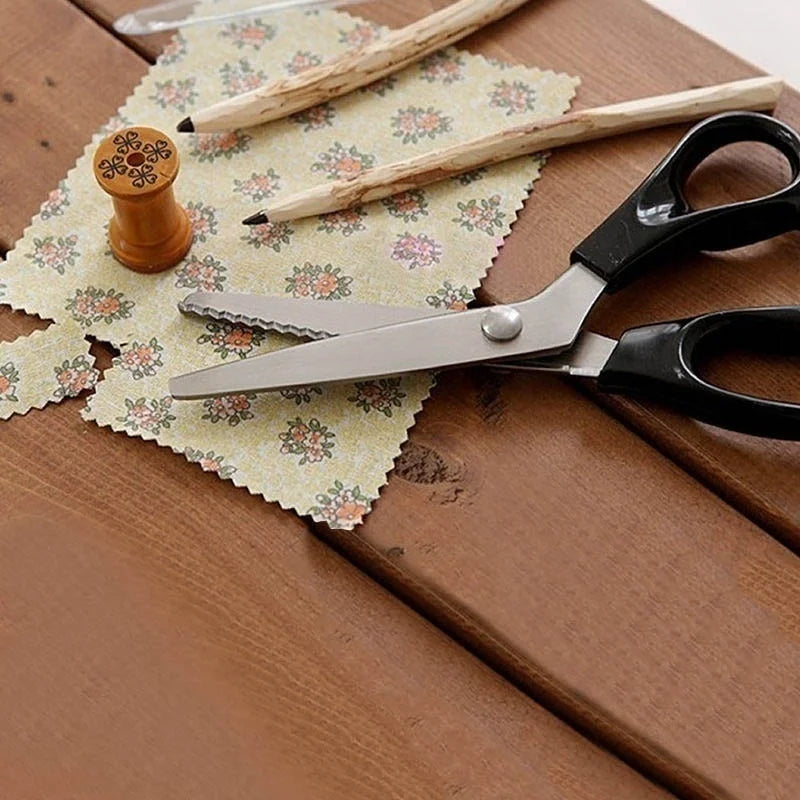 A pair of professional scissors with scalloped edges rests on a wooden surface next to a pencil and patterned paper.