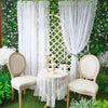 Two vintage floral lace sheer curtains hang in front of a green hedge with white flowers and a small table set for an outdoor