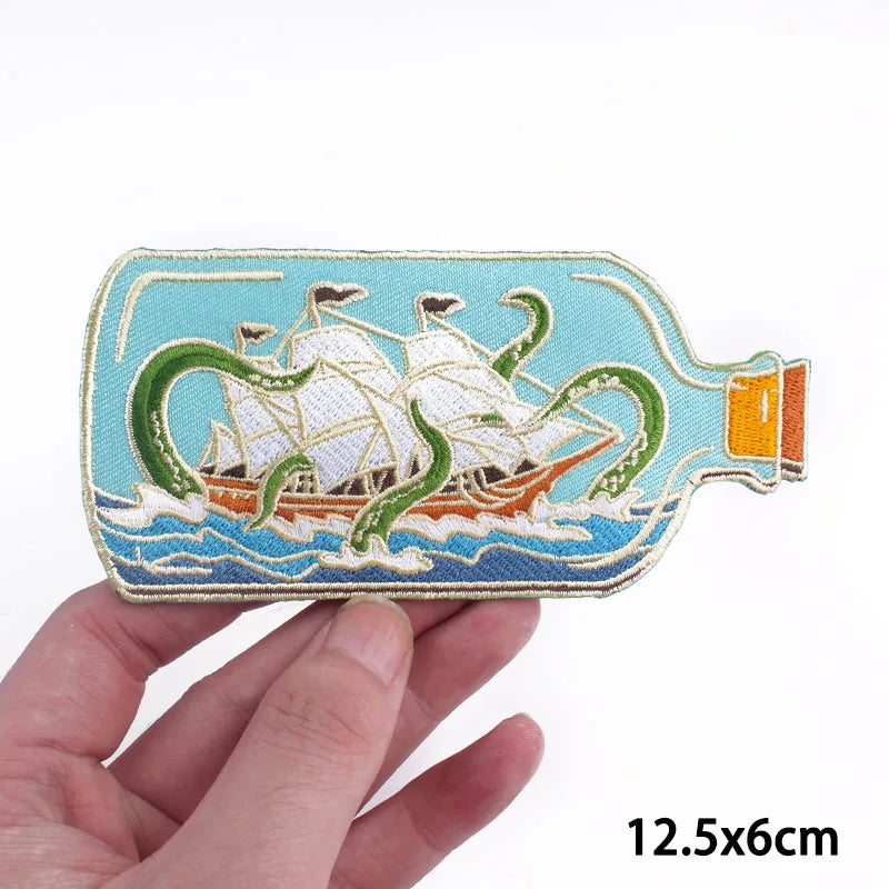 A hand holds a light blue embroidery patch depicting cartoon cats sailing on the water.