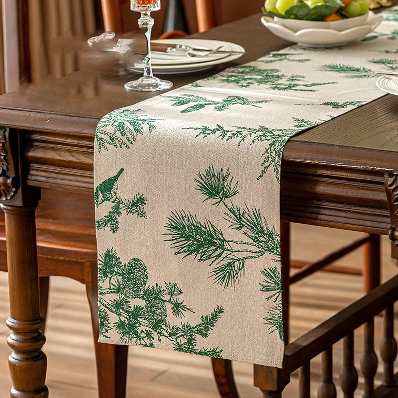 A beige table runner with a green pine branch pattern is placed on a dark wooden dining table.