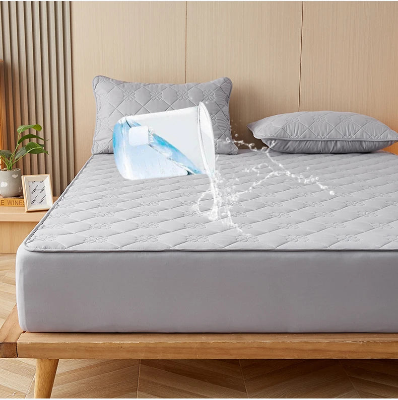 A gray mattress protector is shown on a bed, with water splashing from a glass onto the fabric.