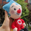A hand holds a soft, blue-haired clown plush toy with a red nose and a red shirt against a blurred green background.